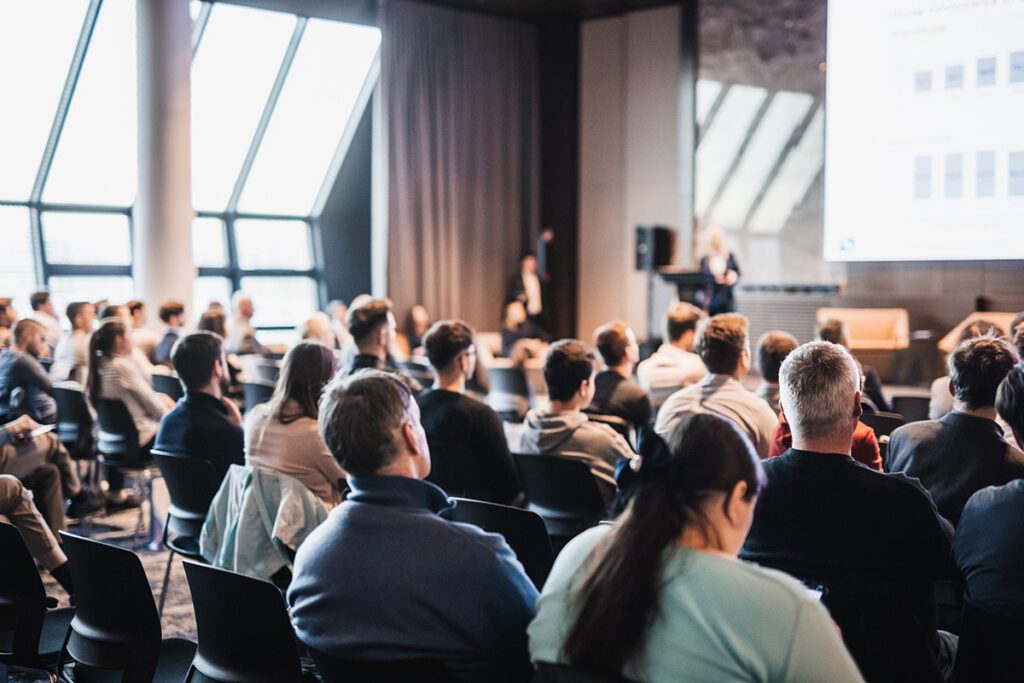 Engaged audience listening to Dr. Belle deliver a keynote speech at a professional conference or speaking engagement, with a presenter visible at the front of the room